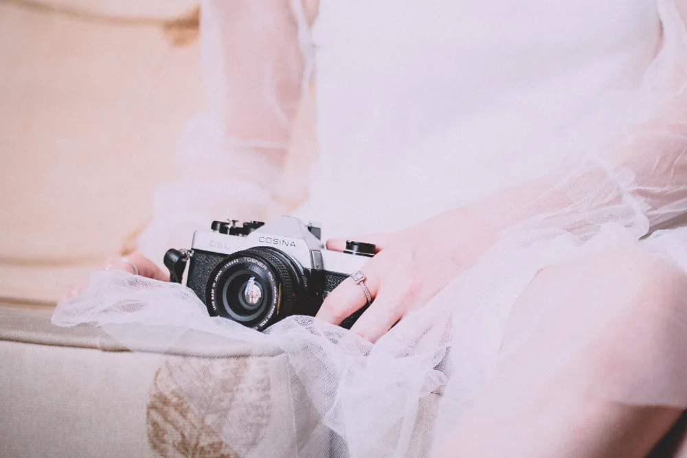 Bride in a sheer white dress holding a vintage film camera on her lap, illustrating wedding photography content on Instagram in the US