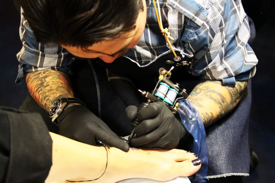 Tattoo artist in black gloves working on a client's foot with a coil tattoo machine, illustrating tattoo art content on Instagram in the US