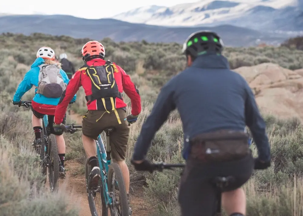 Mountain bikers riding a singletrack trail through sagebrush toward snowy peaks in Colorado, the kind of terrain endurance mountain bike racers train on